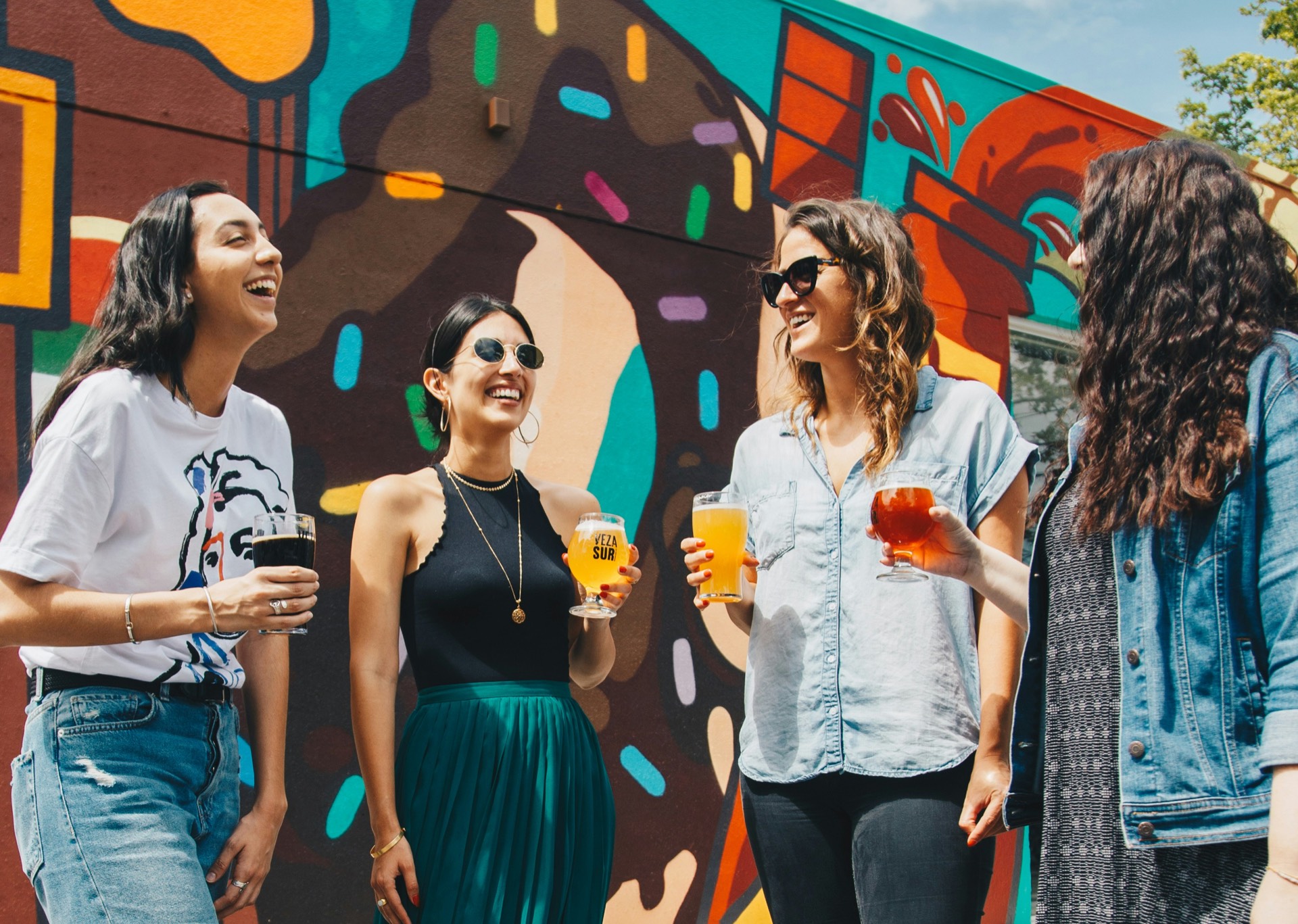 Four friends laughing together over drinks in front of a colourful street mural